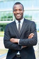 Confident and successful businessman. Handsome young African man in full suit keeping arms crossed and looking at camera while standing outdoors photo