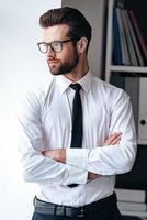 Searching for new solution. Serious young businessman in glasses keeping arms crossed and looking away while standing in office photo