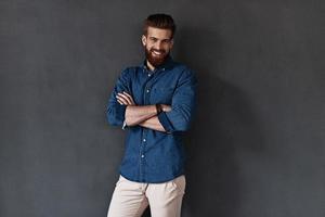 Trendy look. Good looking young man keeping arms crossed and looking at camera while standing against grey background photo