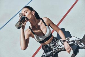 She needs to be hydrated. Top view of young woman in sports clothing drinking water while sitting on the running track outdoors photo