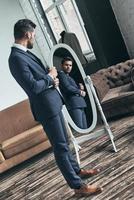 Elegant look. Full length of handsome young man in full suit adjusting his jacket while standing in front of the mirror indoors photo
