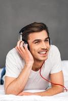 Enjoying good music in bed. Cheerful young man adjusting his headphones and looking away with smile while lying on front in bed against grey background photo