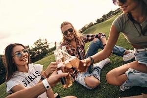 grupo de jóvenes sonrientes con ropa informal brindando con botellas de cerveza mientras disfrutan de un picnic al aire libre