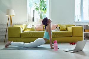 Confident young woman in sports clothing doing stretching exercises with her dog at home photo