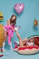 Doing whatever they want. Beautiful young woman removing roller skate from her friend foot and smiling while posing against blue background photo