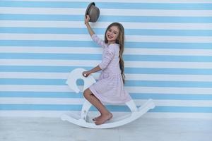 On the way to her dreamland. Cute little girl waving with her hat and looking at camera with smile while sitting on the toy horse photo