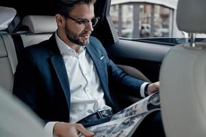 Handsome young man in full suit reading a newspaper while sitting in the car photo