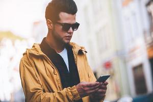 Quick message to friend. Handsome young man holding smart phone and looking at it while standing on the city street photo