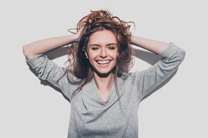 felicidad verdadera. Atractiva joven mujer sonriente cogida de la mano en el cabello y manteniendo los ojos cerrados mientras se enfrenta a un fondo gris foto