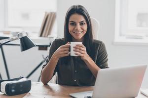 Confident and inspired. Confident young woman in smart casual wear holding coffee cup and looking at camera with smile while sitting at her working place in office photo