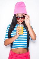 Enjoying fresh juice. Attractive young African woman in funky clothes drinking orange juice and looking at camera while standing against white background photo