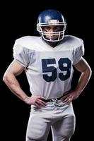 American football player. American football player looking at camera and holding hands on hips while standing against black background photo