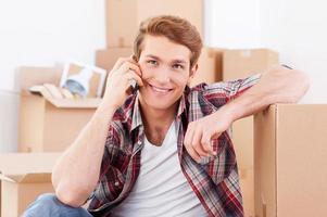 Calling from new house. Handsome young man sitting on the floor and talking on the mobile phone while cardboard boxes laying on the background photo