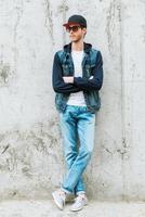Confident style. Handsome young man in headwear keeping arms crossed while standing against concrete wall photo