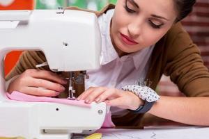 ella es una sastre experta. diseñadora de moda femenina segura de sí misma trabajando en una máquina de coser mientras se sienta en su lugar de trabajo foto