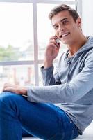 charla despreocupada. vista superior de un joven feliz hablando por teléfono móvil y sonriendo mientras se sienta en el alféizar de la ventana foto