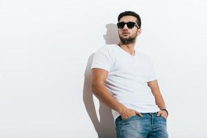 Charming macho. Low angle view of handsome young Indian man in sunglasses holding hands in pockets and looking away while standing against white background photo