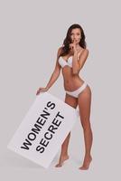 Shhh...  Full length of attractive young woman holding a poster and keeping index finger by her lips while standing against grey background photo