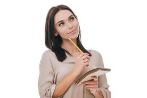 Waiting for inspiration. Thoughtful young woman in smart casual wear holding notebook and pencil while standing against white background photo