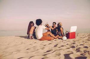 Cheers to friends Cheerful young people spending nice time together while sitting on the beach and drinking beer photo