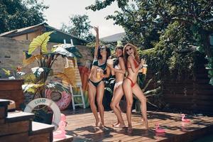 Full length of attractive young women in swimwear having fun with refreshing cocktails and smiling while standing on the poolside outdoors photo