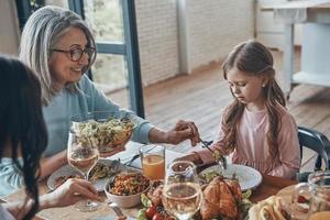 Beautiful senior woman putting salad for little girl while having dinner together photo