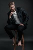 Thoughtful handsome. Full length of stylishly dressed young man looking at camera and holding hand on chin while sitting on chair against grey background photo