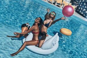 Top view of playful young woman in bikini throwing a ball while floating on a big inflatable swan with her friend photo