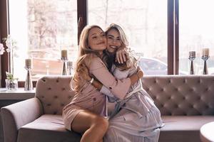 Best friends ever. Attractive young women in elegant dresses embracing and smiling while sitting on the sofa photo