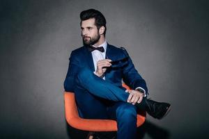 Stylish as ever. Young handsome man in suit holding his sunglasses and looking away while sitting in orange chair against grey background photo