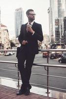 Having a coffee break. Confident young man in full suit holding coffee cup and looking away while standing outdoors with cityscape in the background photo