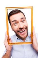 Joyful frame. Handsome young man in shirt holding picture frame in front of his face and smiling while standing against white background photo