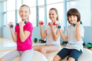 mantener nuestro cuerpo en forma. madre alegre y dos niños haciendo ejercicio con pesas en el club de salud mientras se sientan juntos en las pelotas de fitness foto