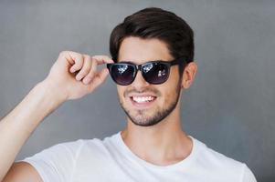 sincero guapo. un joven apuesto que ajusta los anteojos y sonríe mientras se enfrenta a un fondo gris foto