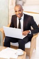 Businessman with laptop. Cheerful young African businessman using his laptop and smiling at camera while sitting at the restaurant photo