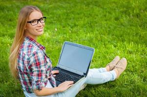 Surfing the net in nature. Beautiful young female student working on laptop and looking over shoulder while sitting on the grass in park photo