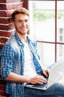 Man with laptop. Top view of handsome young man working on laptop while sitting on the window sill photo