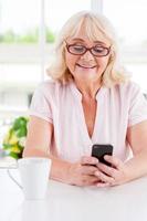 Message from her nearest. Cheerful senior woman looking at her mobile phone and smiling while sitting at the table photo