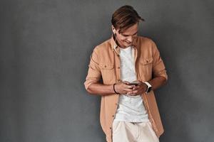 Searching for the best song. Handsome young man holding a smart phone and smiling while standing against grey background photo
