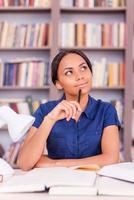 sintiéndose segura de sus exámenes finales. joven negra segura de sí misma tocándose la barbilla con un bolígrafo y mirando hacia otro lado mientras se sienta en el escritorio de la biblioteca foto