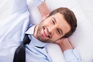 feliz día soñador. vista superior de un apuesto joven con camisa y corbata cogidos de la mano detrás de la cabeza y sonriendo mientras está acostado en la cama foto