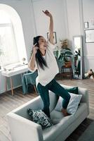 Superstar inside her. Full length of attractive young woman wearing headphones and smiling while dancing on sofa at home photo