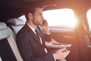 siempre disponible. un joven apuesto con traje completo hablando por teléfono inteligente mientras está sentado en el auto foto