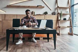 Young and successful. Handsome young man using his smart phone while sitting in the creative working space photo