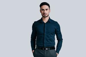 Confidence and charisma. Good looking young man keeping hands in pockets and looking at camera while standing against grey background photo