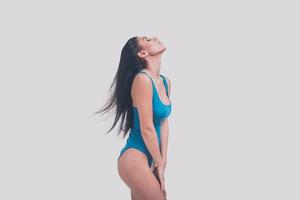 She is gorgeous. Side view of attractive young woman in blue swimsuit keeping eyes closed while standing against grey background photo