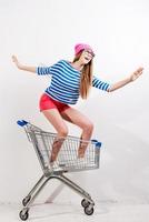Extreme shopping. Playful young woman in headwear and glasses riding in shopping cart against grey background photo