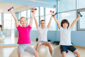 disfrutando de un estilo de vida saludable. madre alegre y dos niños haciendo ejercicio con pesas en el club de salud mientras se sientan en las pelotas de fitness foto