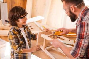 esto se va a ver increíble. niño alegre ayudando a su padre a tosar una silla de madera en el taller foto