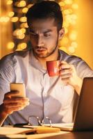 Having no time for break. Confident young man looking at his smart phone and holding coffee cup while sitting at his working place at night time with Christmas lights in the background photo